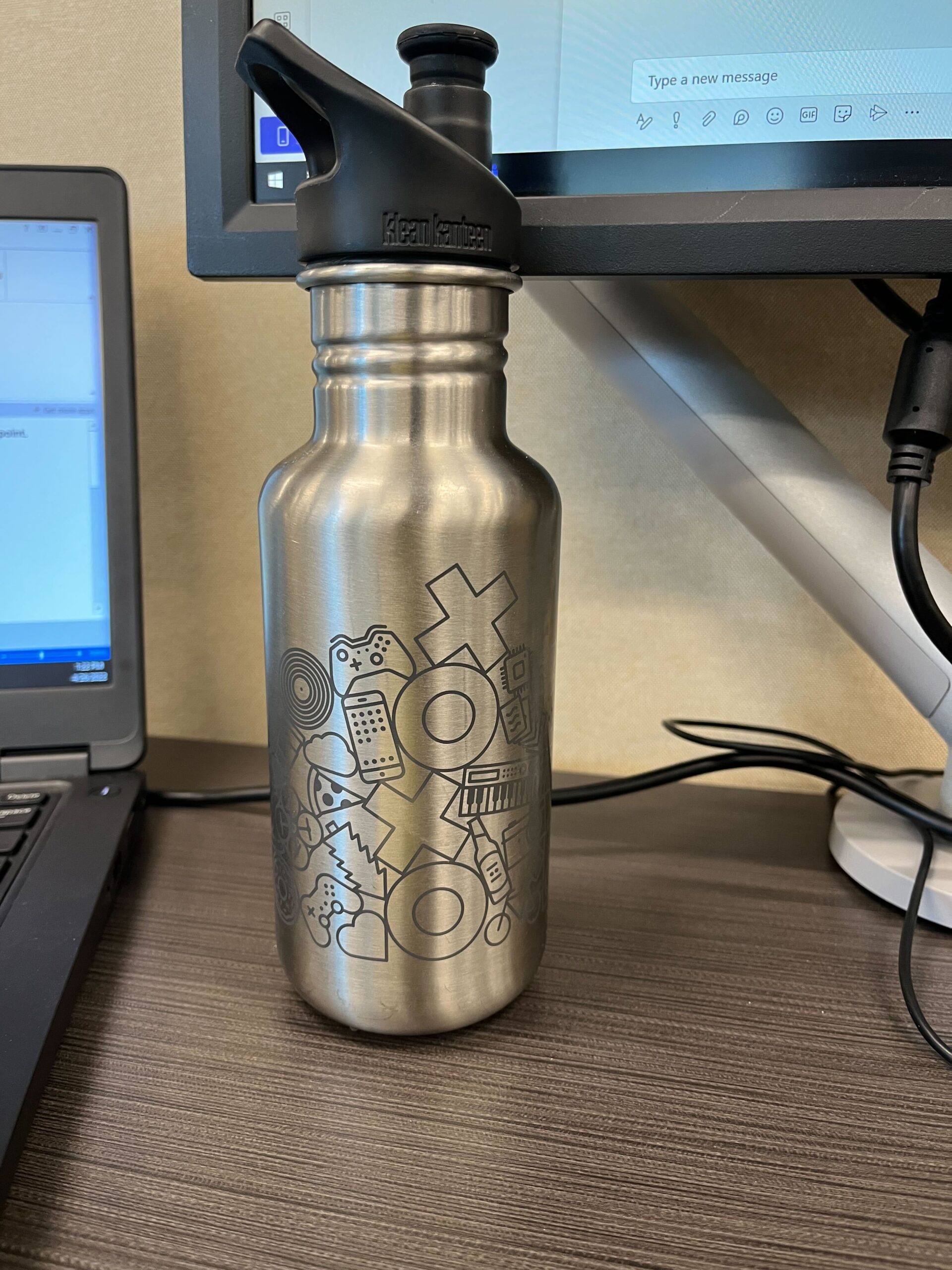 A picture of a work desk. To the left there's a laptop; to the upper right is the lower corner of a monitor. In the middle is a stainless steel water bottle with the logo of the XOXO Festival printed on it.