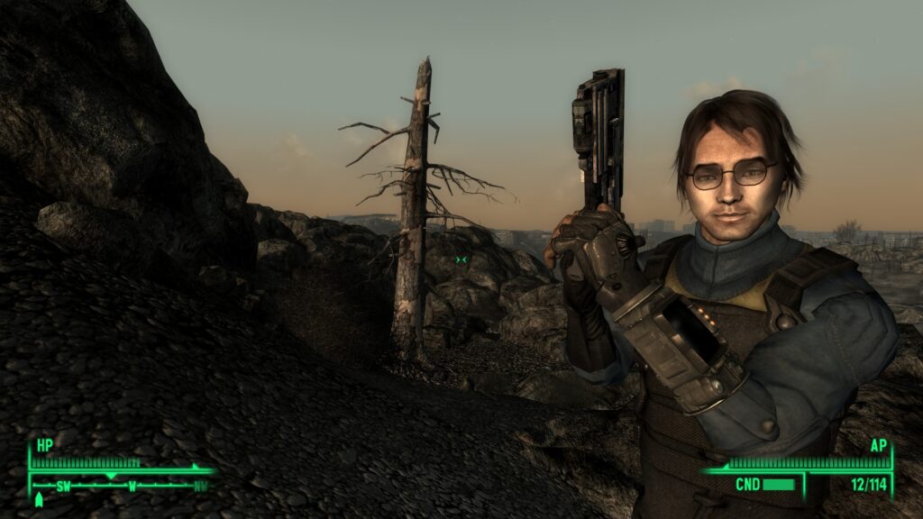 Screenshot from the game Fallout 3. A young brown-haired white man stands on a ledge overlooking a desolate ruin. He holds a 10mm pistol up with both hands and is wearing an armored Vault Suit.