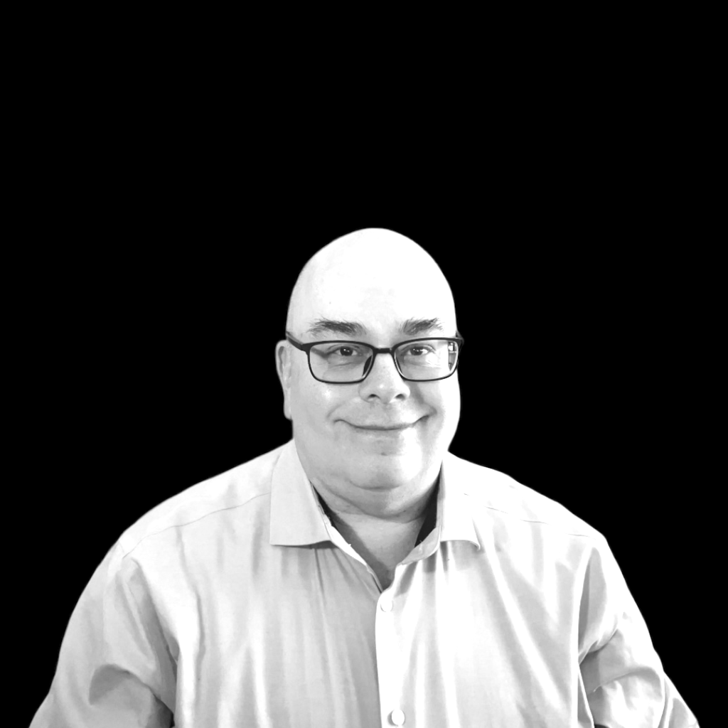 Brian Moon, a bald white older man, smiling. He's wearing eyeglasses, and a buttoned shirt. The background is a corner office on an upper floor, with an open laptop on a table and a potted tree. The picture is in black and white.