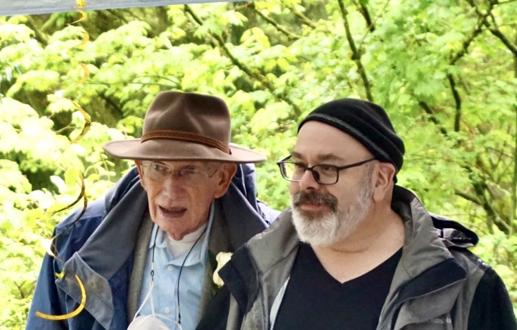 Don, a 90 year old white man wearing a broad-brimmed hat and a coat, smiling, next to a middle-aged white man with a black stocking cap, glasses, and a coat, outside, with a background of green leaves behind them.