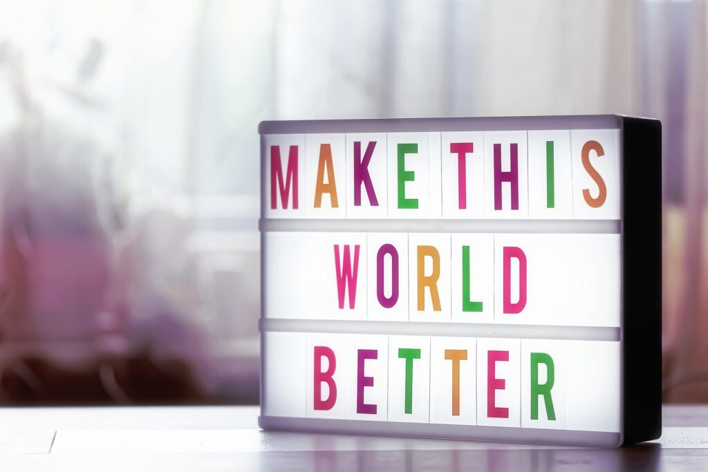 A sign, lit up from the inside, that says "Make This World Better" in friendly multi-colored letters sits on a shelf, with a blurry background overlit by sunshine.