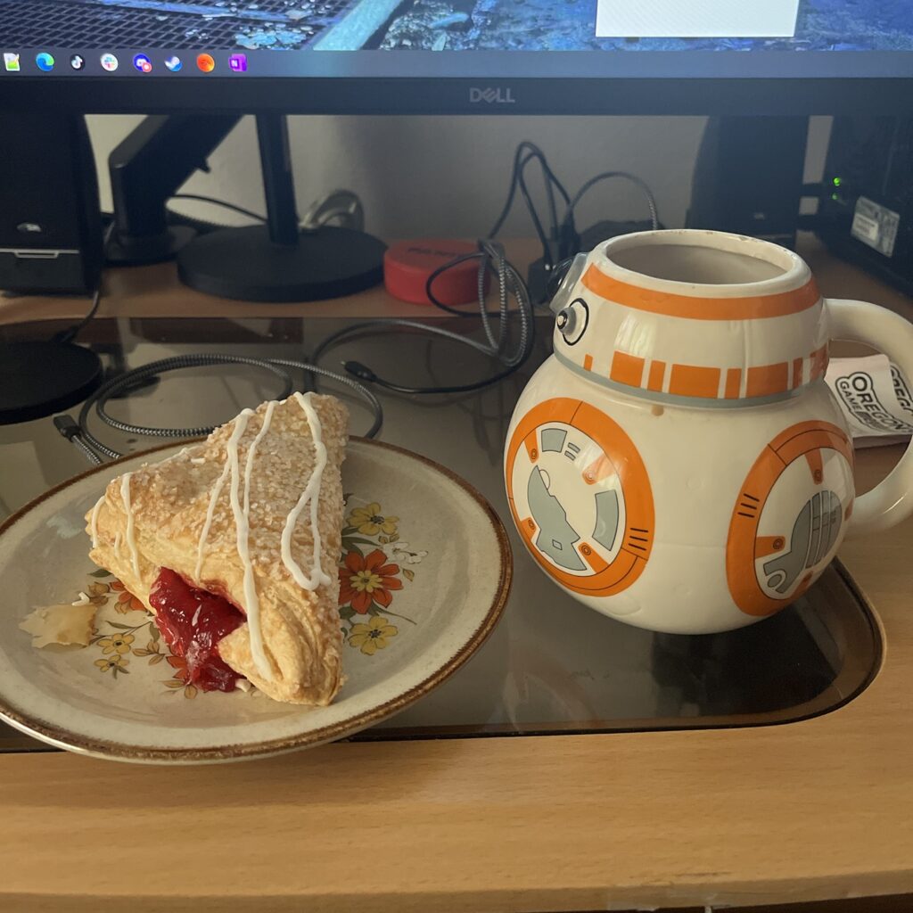 A cherry turnover on a plate next to a coffee mug in the shape of BB-8 from Star Wars. Both are sitting on a computer desk. In the background on the desk are a pair of computer speakers, the arms for a computer monitor, a USB hub with cables coming out of it, and a stack of Oregon Megabucks lottery tickets.