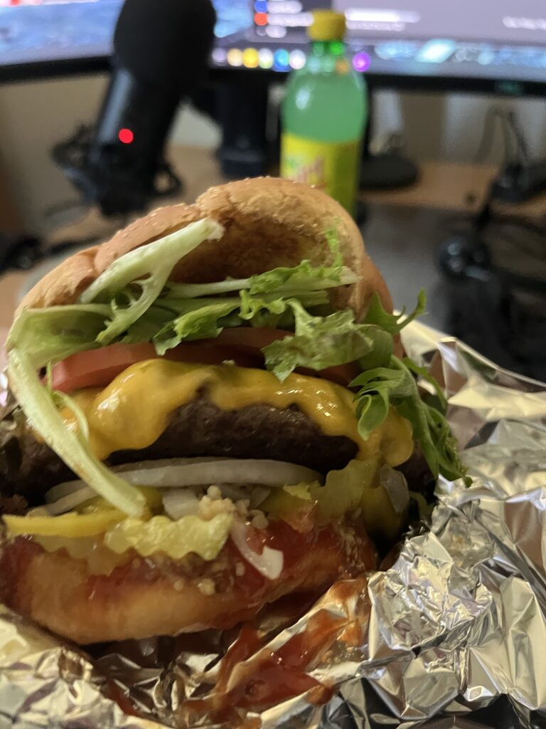 Close up of a homemade cheeseburger; lettuce, tomato slices, white onion slices, pickles and banana peppers, along with catsup, spill out of it onto the aluminum foil that wraps it up. In the background is a Yeti microphone, a bottle of Squirt soda, and the bottom edge of two computer monitors. This person is eating at their desk.