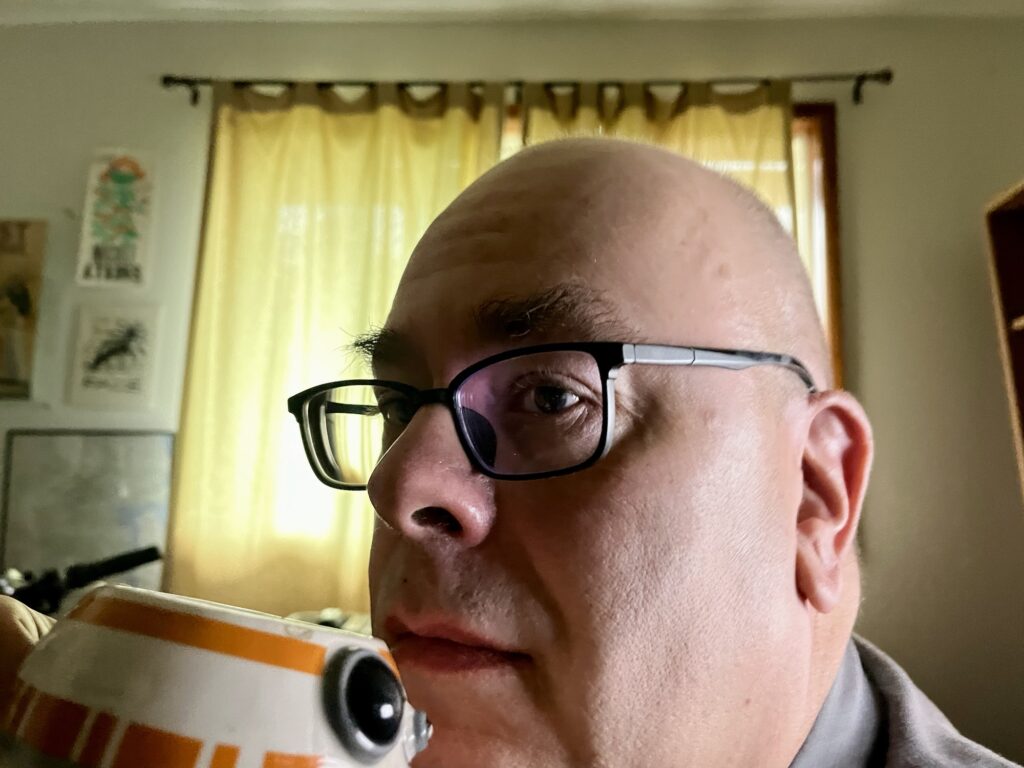 Close up of the author, a bald middle aged white guy wearing black framed glasses. He's shaved today. He's holding up a BB-8 shaped coffee mug. Behind him, yellow curtains are backlit by the sunshine outside. Some framed and unframed prints hang on the wall next to the window.