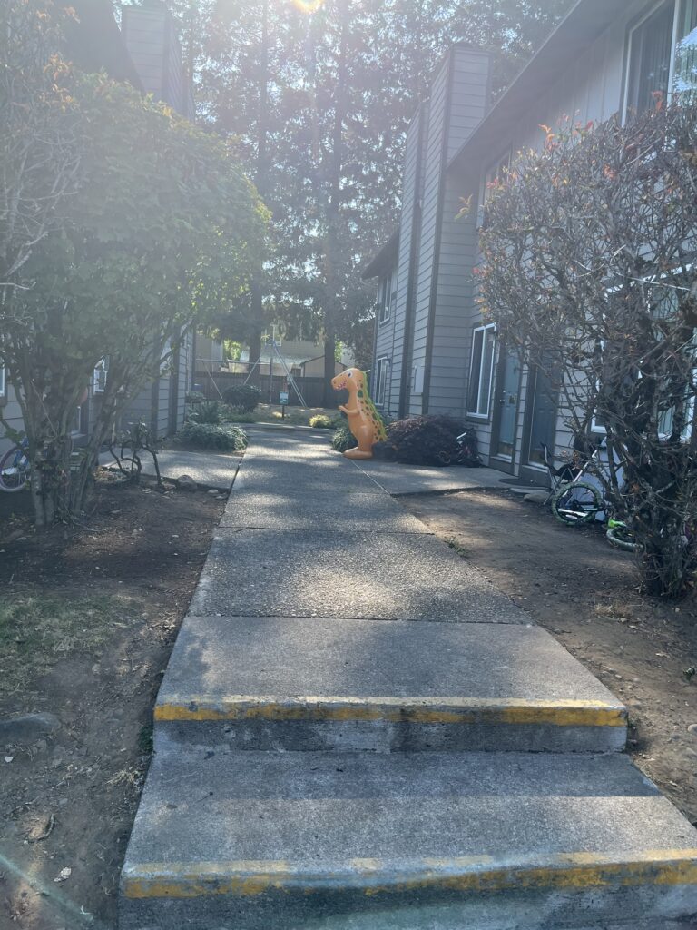 Picture, somewhat faded from being taken facing the sun, of a stair and sidewalk that leads between two rows of townhouses. On the sidewalk, in the middle distance, is a five foot tall inflatable orange dinosaur.