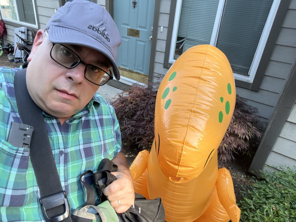 Selfie of the author, a middle-aged white man wearing glasses, an Adidas baseball cap, and a green plaid shirt, standing in front of an orange inflatable dinosaur. Apartment windows and doors are in the background.