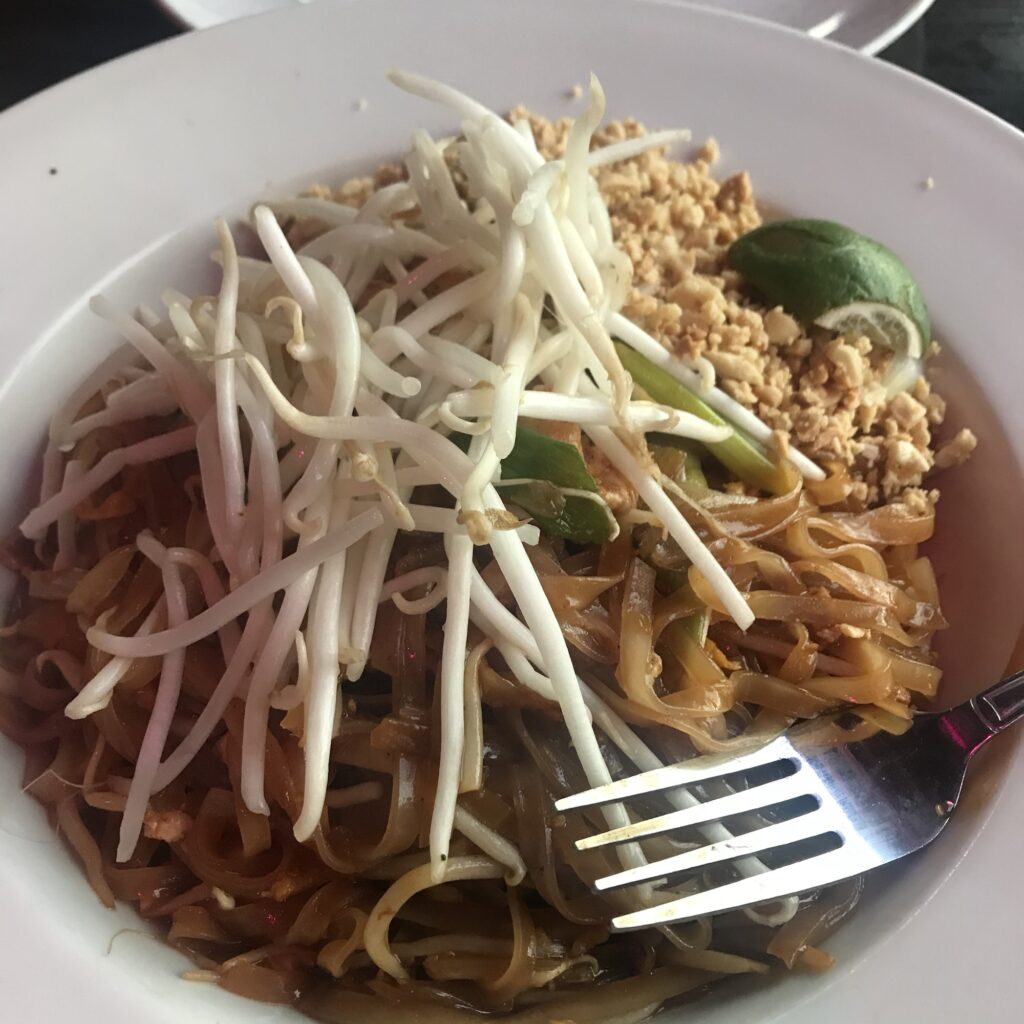 Close up of a white bowl filled with brown pad Thai noodles, with bean sprouts, ground peanuts, green onions, and a slice of lime.