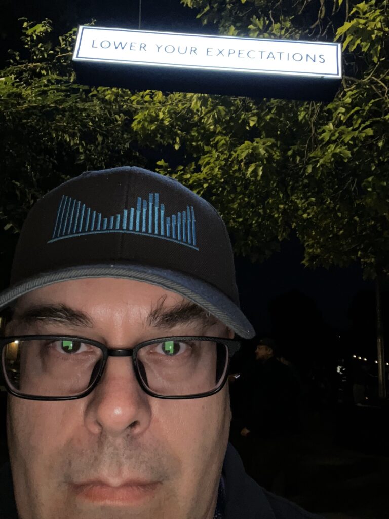 Close-up selfie of the author, a white man wearing glasses and a "The Rebound" baseball cap, from the nose up. A lit sign hangs above him that reads, in simple sans-serif font, "Lower your expectations". It's night time, and the trees in the background are lit up by off-screen lights.